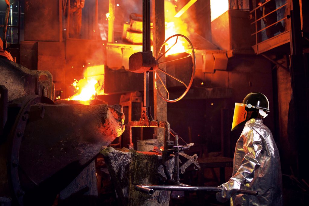 Trabalhador, protegido por EPIs Protenge capa aluminizada e protetor facial, observando o caminho do aço líquido em siderúrgica